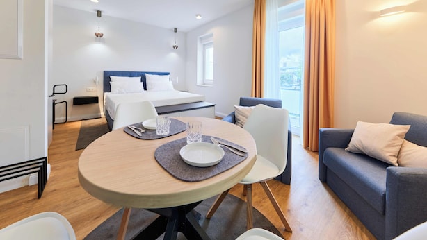 Ein modernes Hotelzimmer mit einem Bett, einem Esstisch mit Stühlen, einer Couch und einem großen Fenster mit Vorhängen.