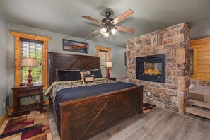 A bedroom with a stone fireplace, a bed with a headboard, a ceiling fan, and a window with blinds. - Mountain High (Gatlinburg)