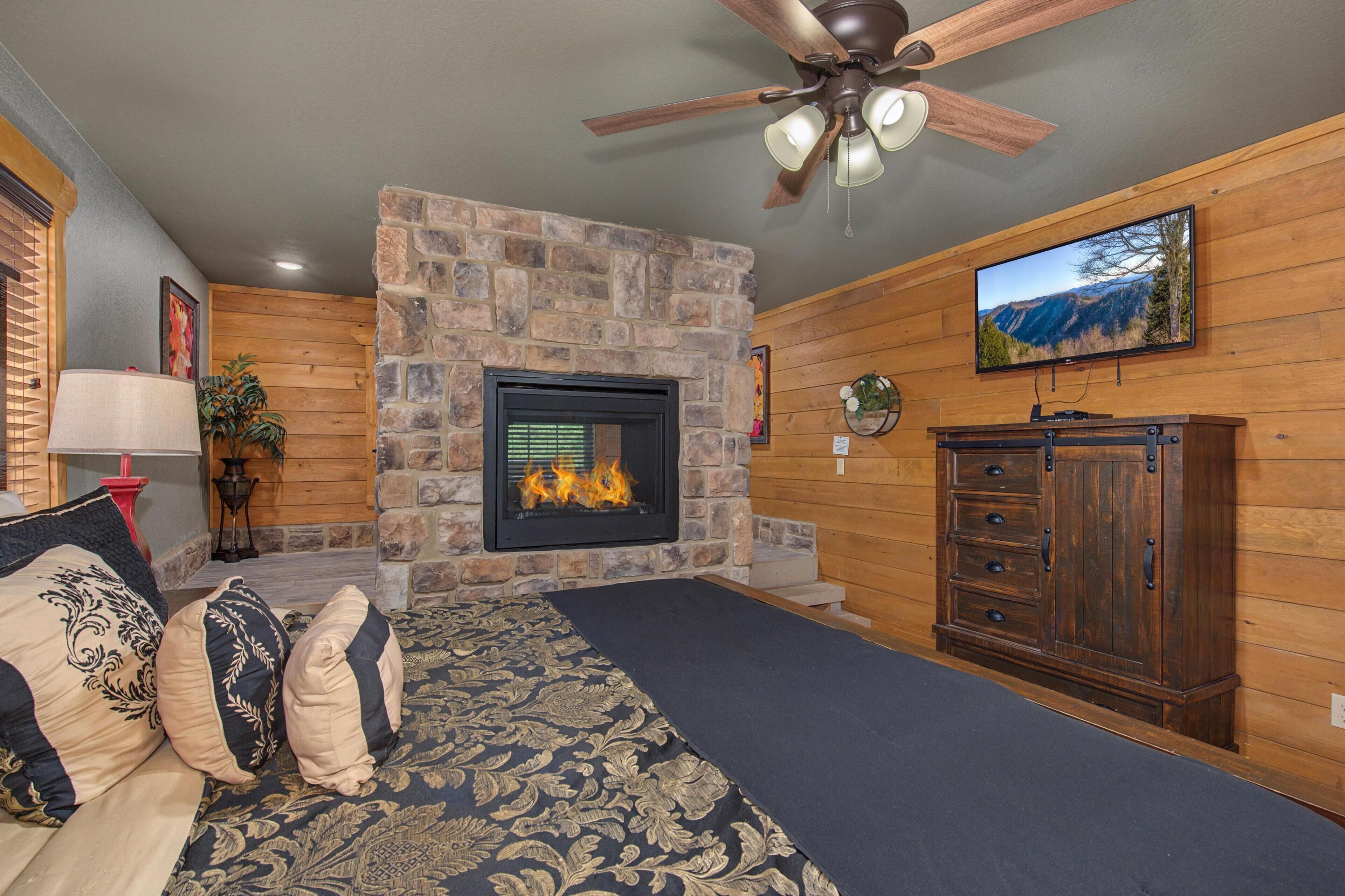 A room with a stone fireplace, a ceiling fan, a television mounted on the wall, and a wooden cabinet.