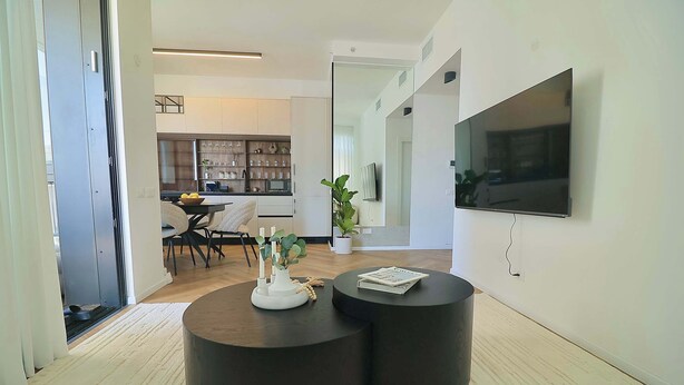 Uma sala de estar moderna com uma TV de tela plana, duas mesas de centro pretas, uma área de jantar com mesa e cadeiras, e uma cozinha ao fundo.