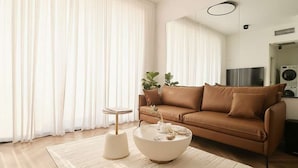 A modern living room with a brown leather sofa, a round coffee table, and a large window with sheer curtains. - High-End 1BR in Secure Tower | Parking | Mamak (Tel Aviv-Yafo)