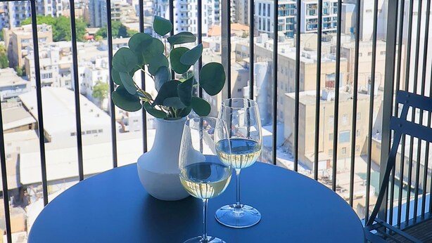 A balcony with a table, two wine glasses, and a potted plant. - High-End 1BR in Secure Tower | Parking | Mamak (Tel Aviv-Yafo)