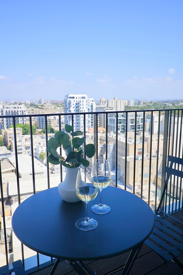 A balcony with a table, two wine glasses, and a potted plant. - High-End 1BR in Secure Tower | Parking | Mamak (Tel Aviv-Yafo)