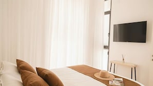 A bedroom with a bed, a television, a bedside table, and a window with curtains. - High-End 1BR in Secure Tower | Parking | Mamak (Tel Aviv-Yafo)