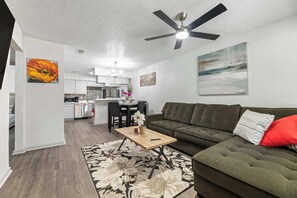A living room with a sofa, a coffee table, and a rug. There is a dining area with a table and chairs. A ceiling fan is visible. - Modern Arlington Escape • Stadium 8 Min Away (Arlington)