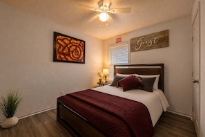 A bedroom with a bed, a ceiling fan, a wall-mounted artwork, a bedside table with a lamp, and a sign on the wall. - Steps from AT&T Stadium | Parking (Arlington)