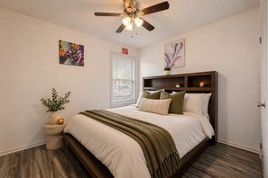 A bedroom with a bed, a ceiling fan, a window with blinds, an exit sign, and a wall-mounted shelf with a plant. - AT&T Stadium Getaway | Parking (Arlington)