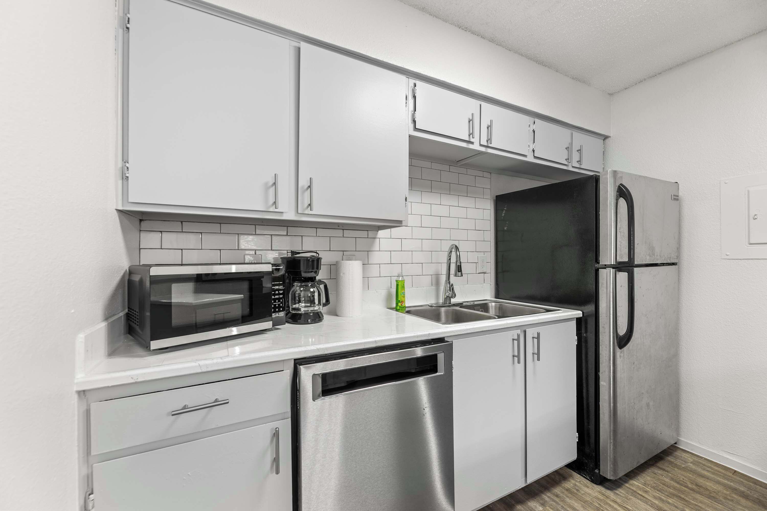A modern kitchen with white cabinets, a stainless steel dishwasher, a microwave, a coffee maker, and a refrigerator.
