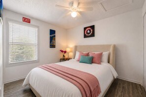 A hotel room with a bed, a ceiling fan, a nightstand with a lamp, a painting, and an exit sign. - City Comfort • 2BR • Stadium Within Walking (Arlington)