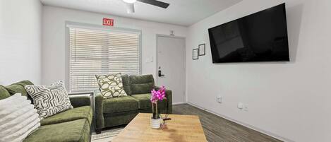 Un salon avec un canapé vert, une table basse en bois, un ventilateur de plafond, un téléviseur à écran plat et un panneau de signalisation indiquant la sortie.