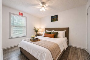 Ein Schlafzimmer mit einem Bett, einem Nachttisch, einer Lampe, einem Deckenventilator, einem Fenster mit Jalousien und einem Fluchtwegschild.