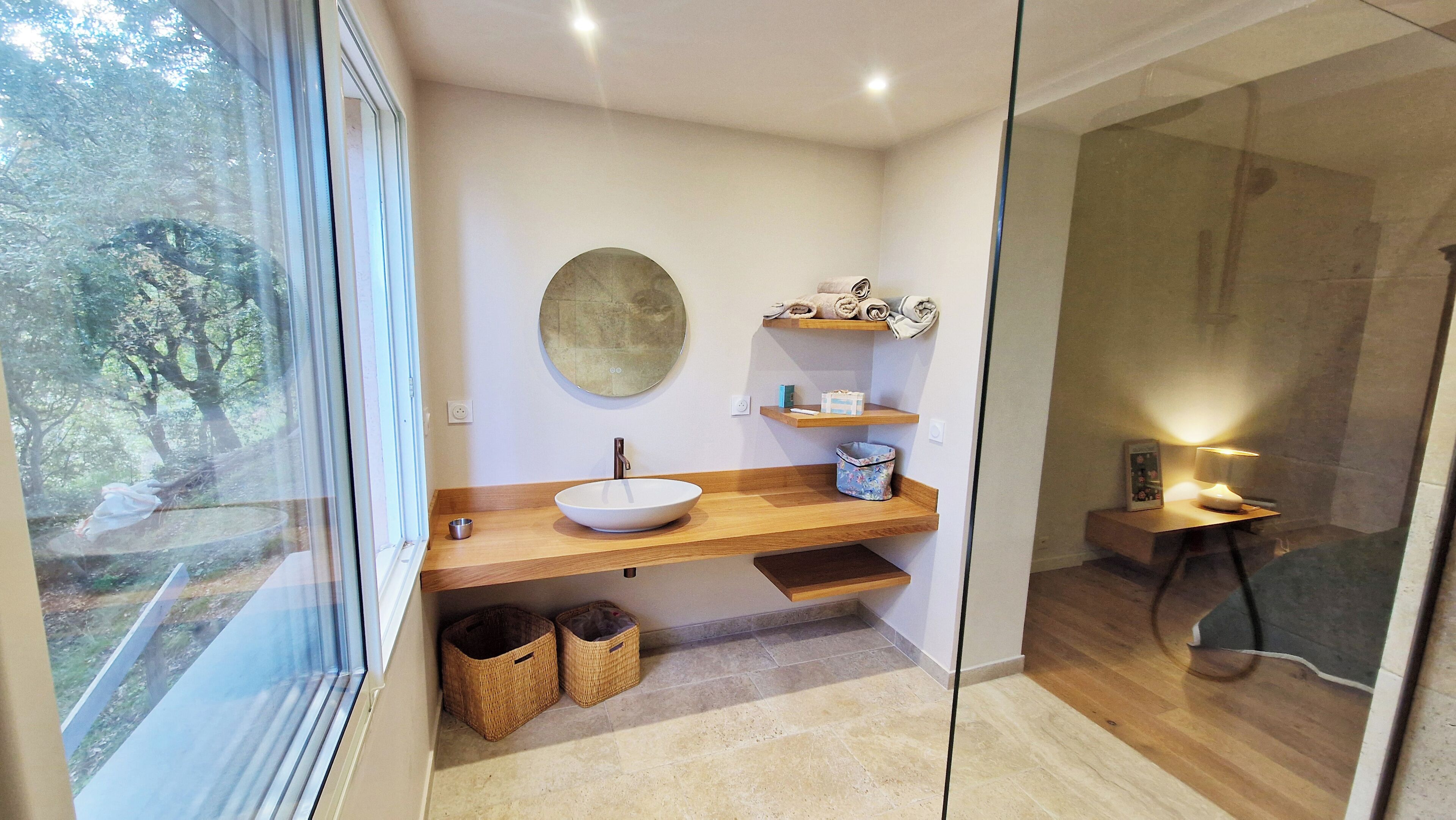 Een moderne badkamer met een groot raam, een ronde spiegel, een houten wastafelmeubel en een klein plankje met handdoeken.