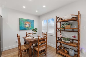 A dining area with a wooden table and chairs, a wall-mounted shelf with decorative items, and a painting on the wall. - Relaxing King Suite With Poolside Patio & Partial Ocean Views (New Smyrna Beach)
