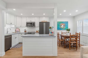 A modern kitchen with white cabinetry, a central island, a dining area with wooden chairs, and a colorful painting on the wall. - Relaxing King Suite With Poolside Patio & Partial Ocean Views (New Smyrna Beach)