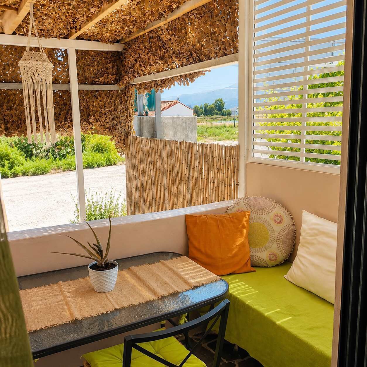 Ein Zimmer mit Blick auf Grünflächen, ein Tisch mit Pflanze, eine gepolsterte Sitzgelegenheit und ein Fenster mit Jalousien.