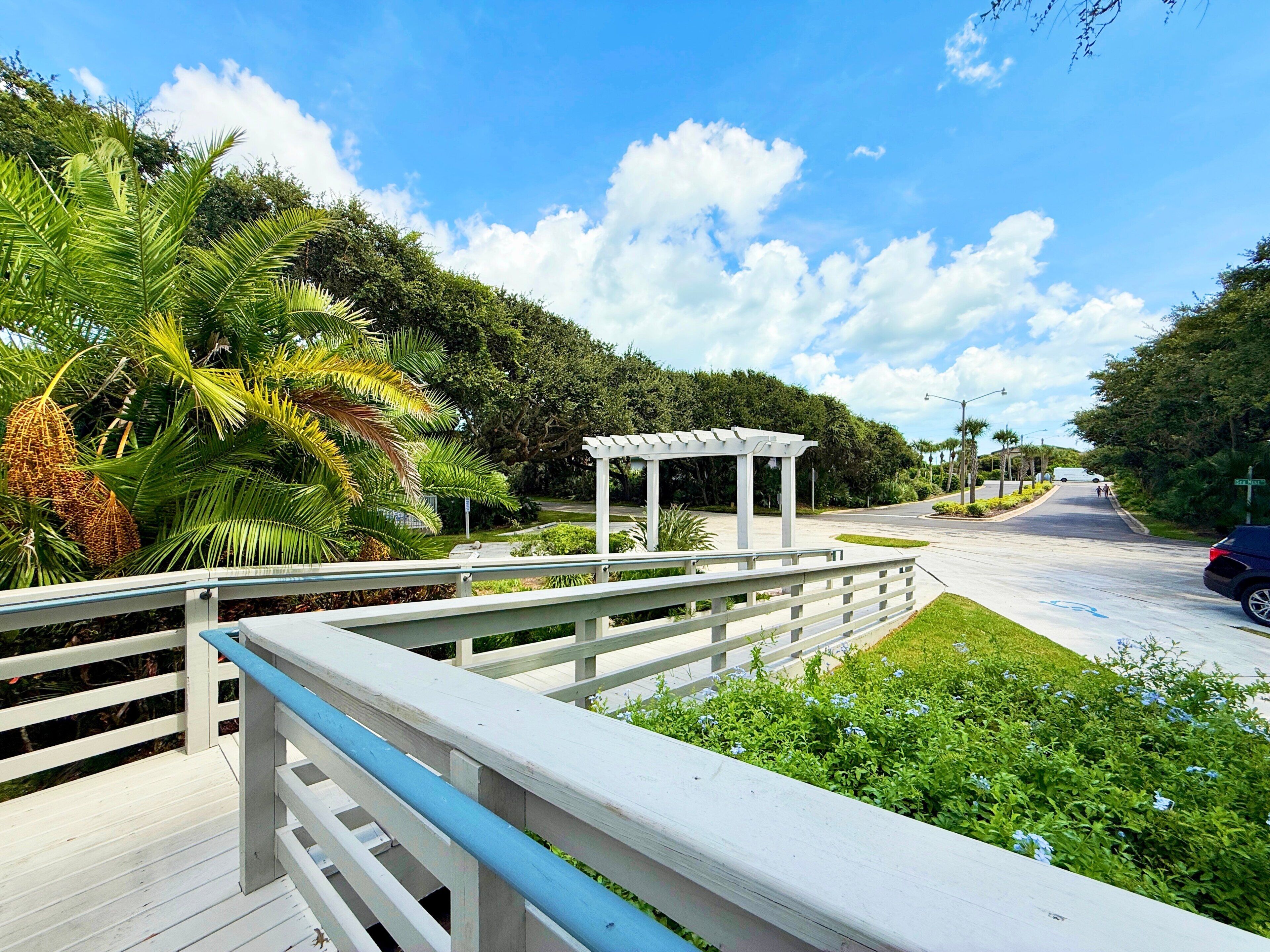 A wooden bridge with a white pergola, surrounded by lush greenery and palm trees.