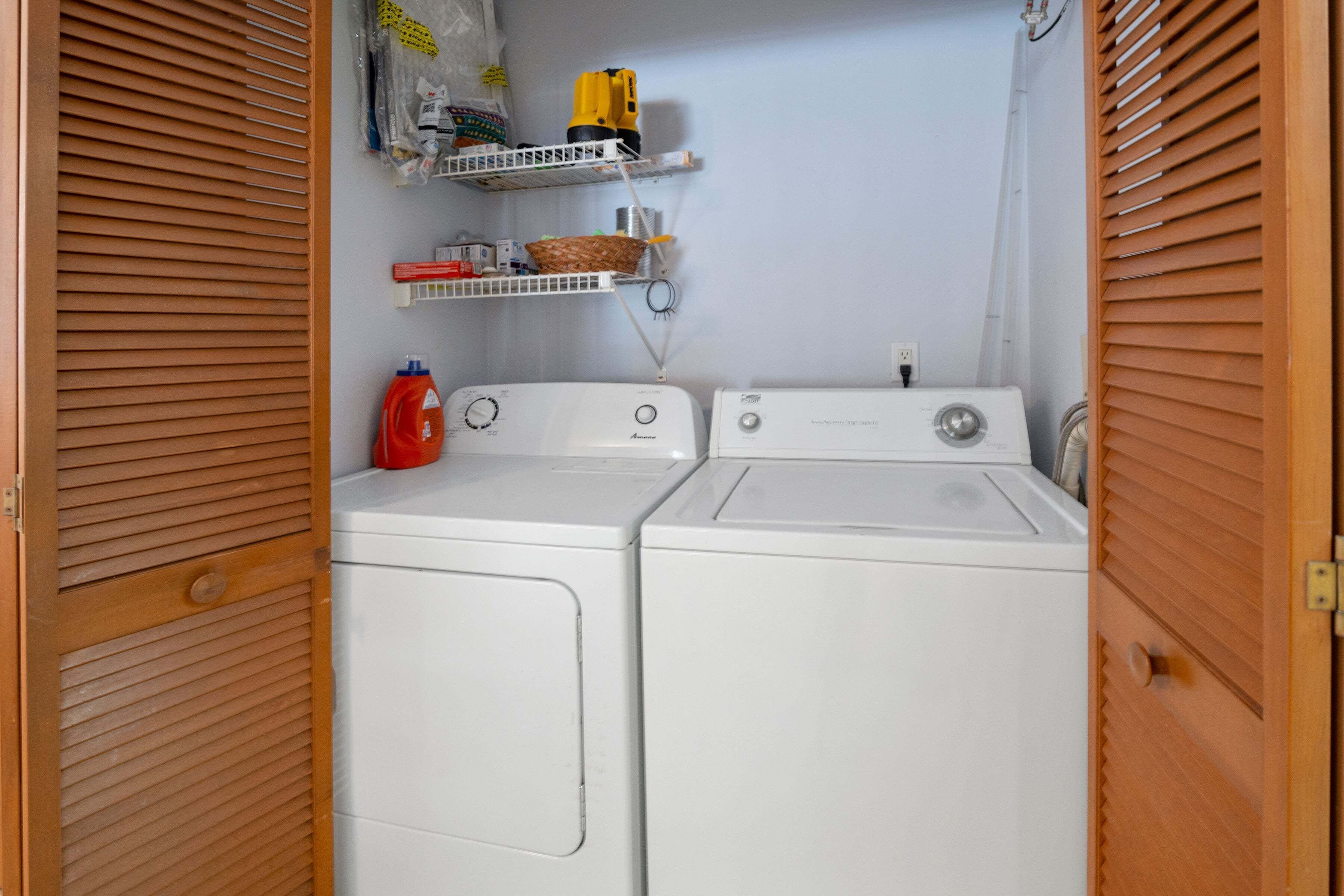 Une buanderie avec deux machines à laver blanches, une bouteille de lessive rouge et des étagères au-dessus contenant divers objets, notamment un outil électrique jaune et un panier.