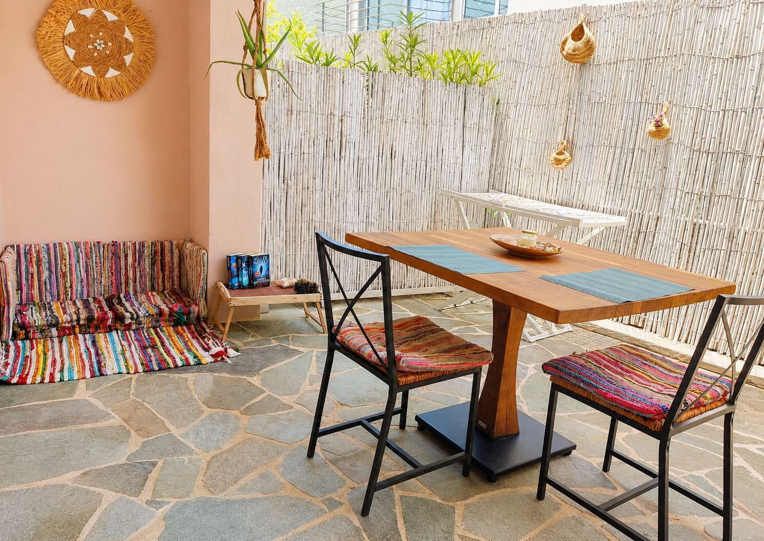 A stone-floored patio with a wooden table and chairs, a woven bench, and a bamboo wall.