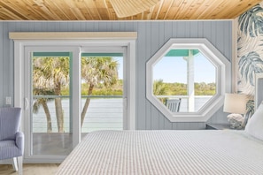 A bedroom with a bed, a chair, a window, and a view of palm trees. - La De Dah (Edgewater)