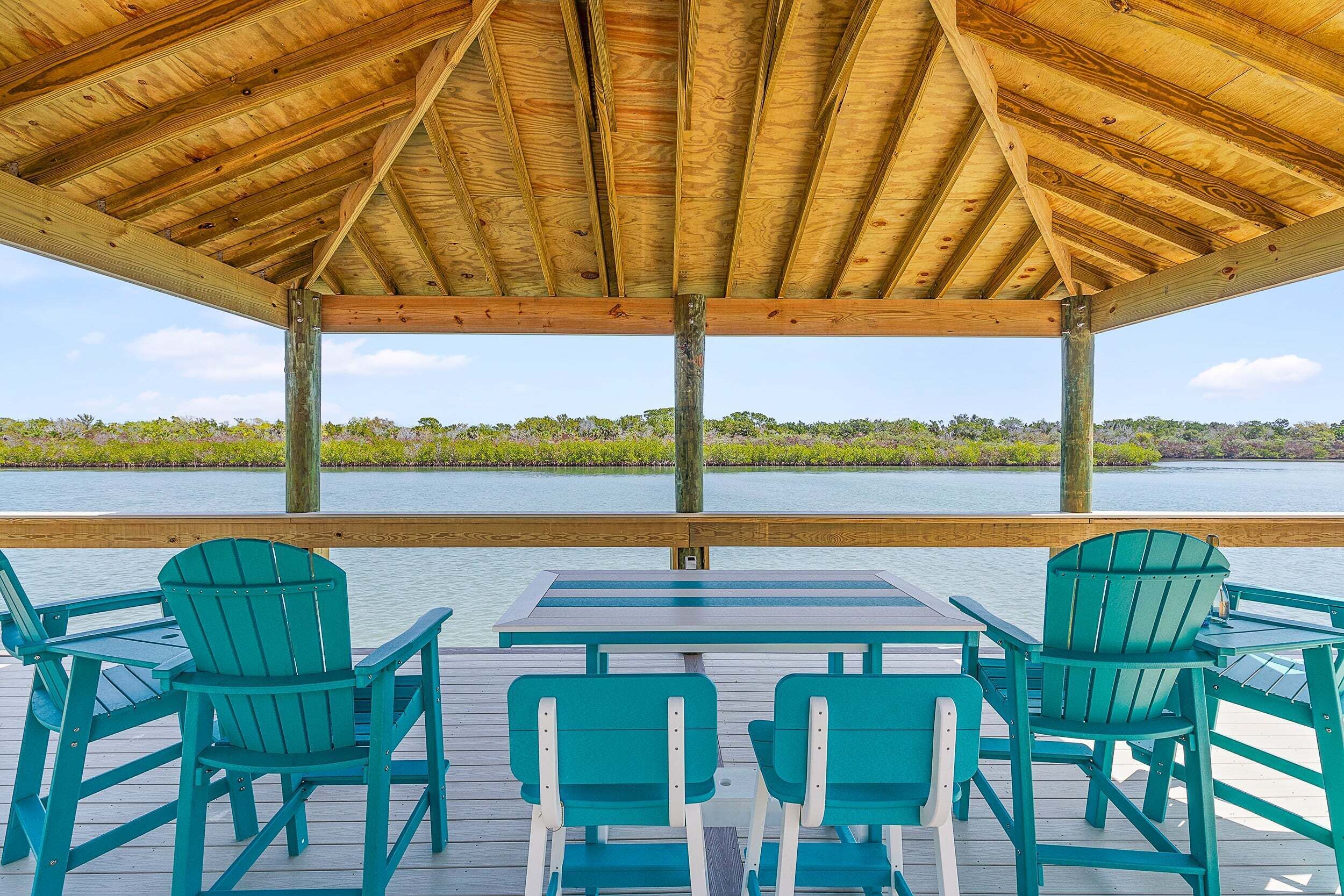 A covered deck with a view of a body of water, wooden beams, and green chairs.