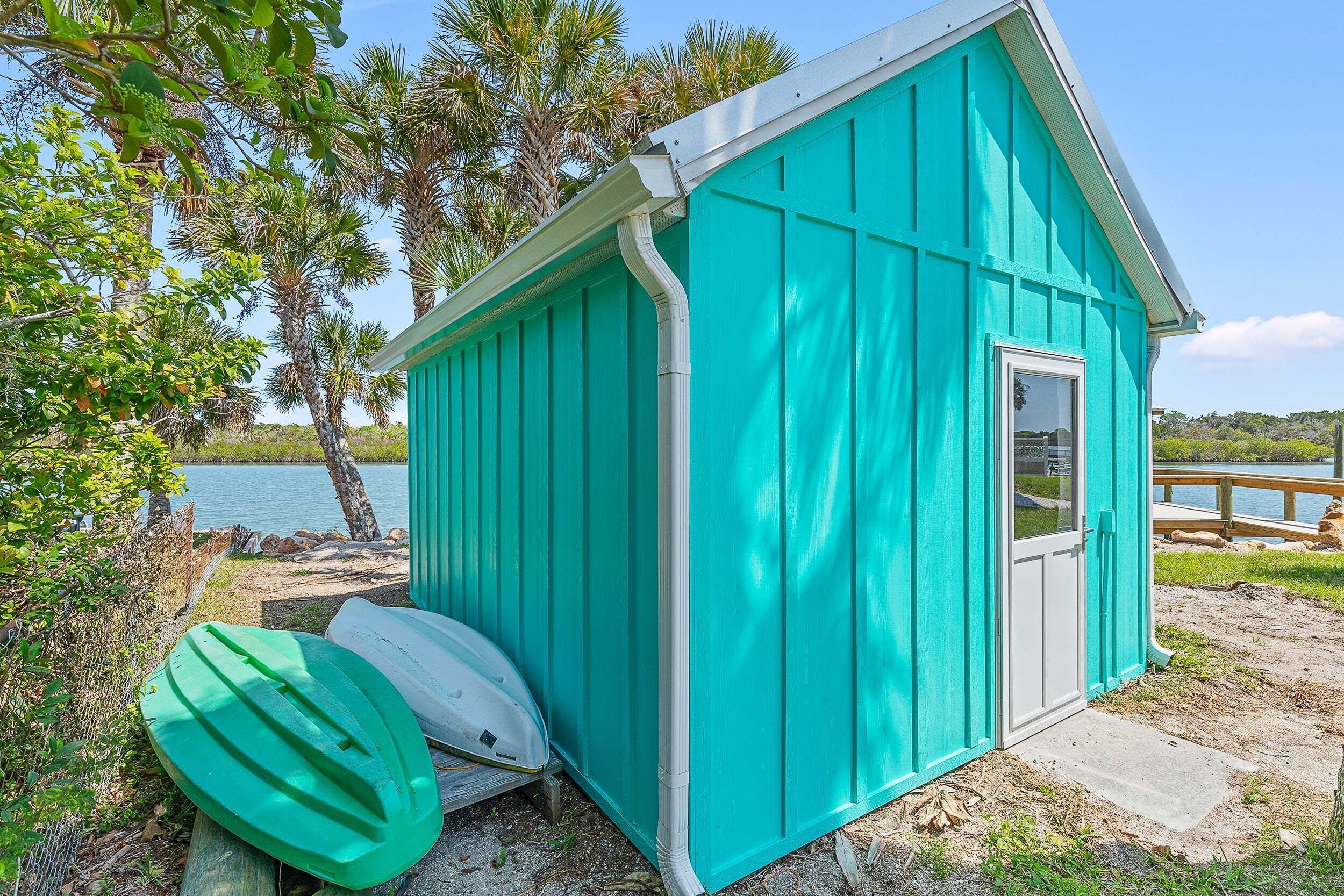 A small, single-story building with a sloped roof, situated by a body of water with palm trees in the background.
