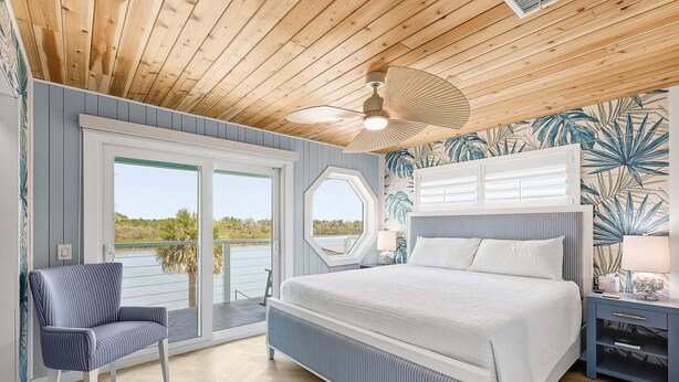 A bedroom with a bed, a chair, a nightstand, and a view through a window. - La De Dah (Edgewater)