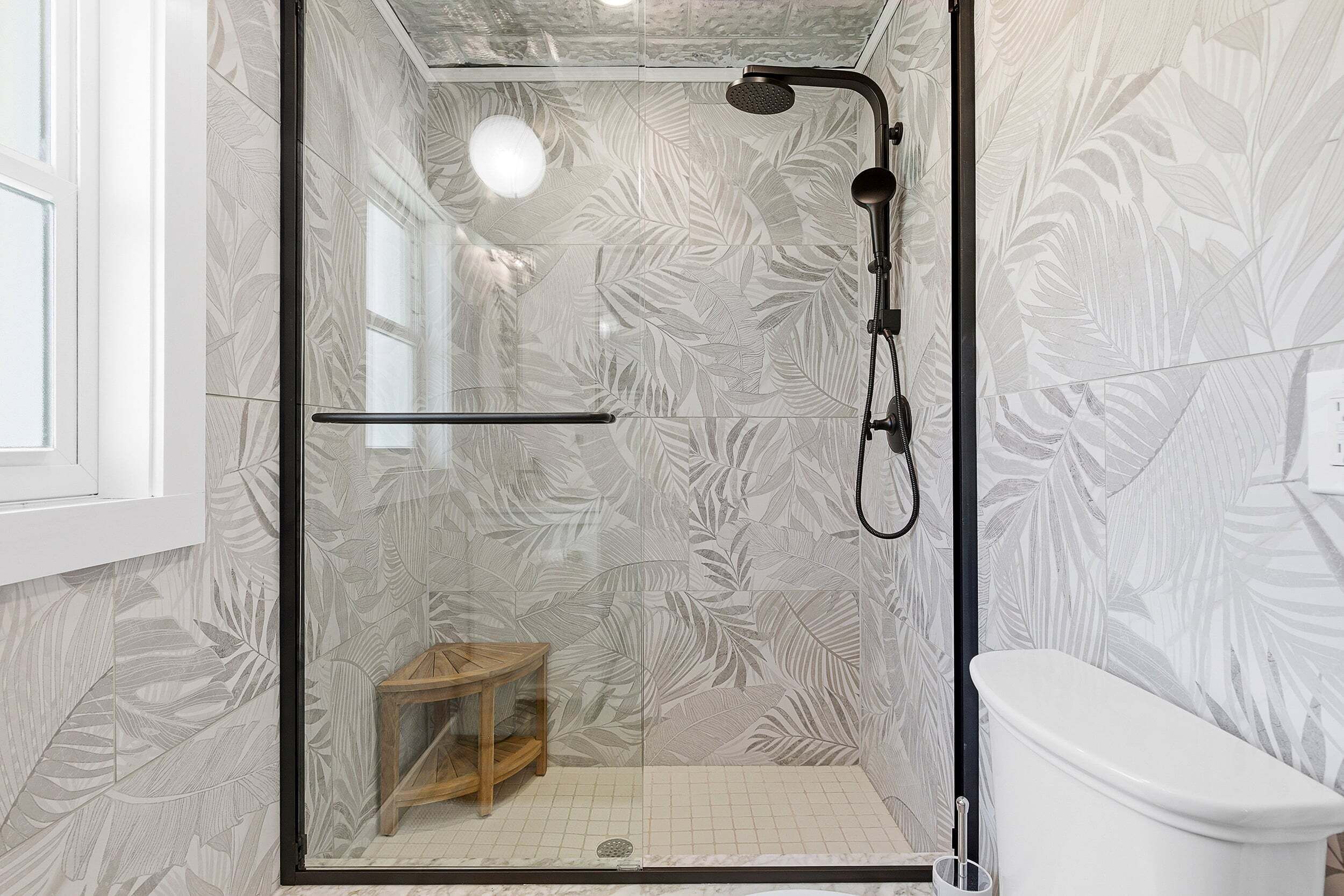A glass-enclosed shower with a wooden stool, a toilet, and a patterned wall.