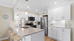 A modern kitchen with white cabinets, granite countertops, stainless steel appliances, and a central island with stools. - Updated Oceanview Retreat With Pools, Pickleball & Direct Beach Access (New Smyrna Beach)