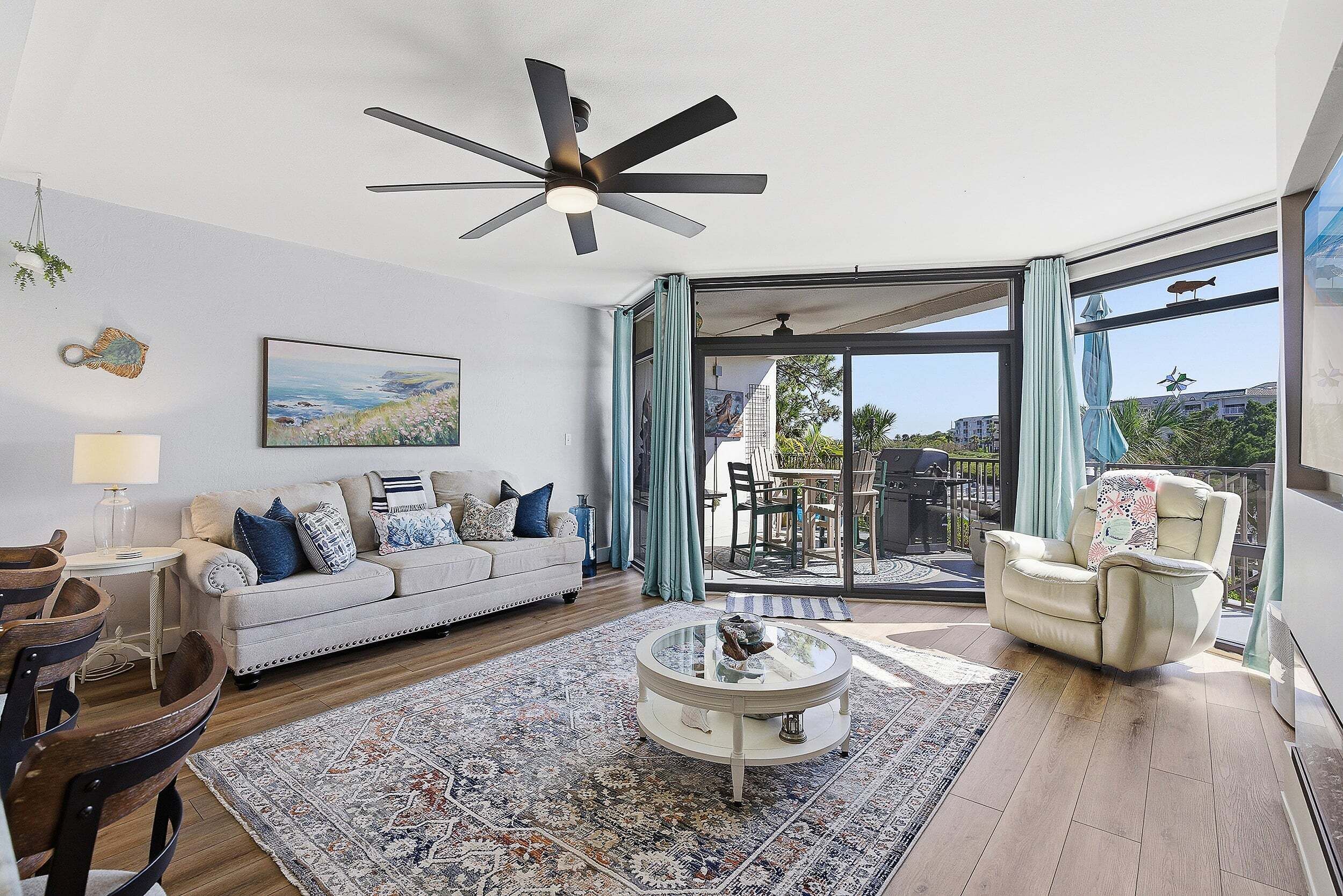 A living room with a sofa, coffee table, and chairs, a ceiling fan, and a large window with a view.