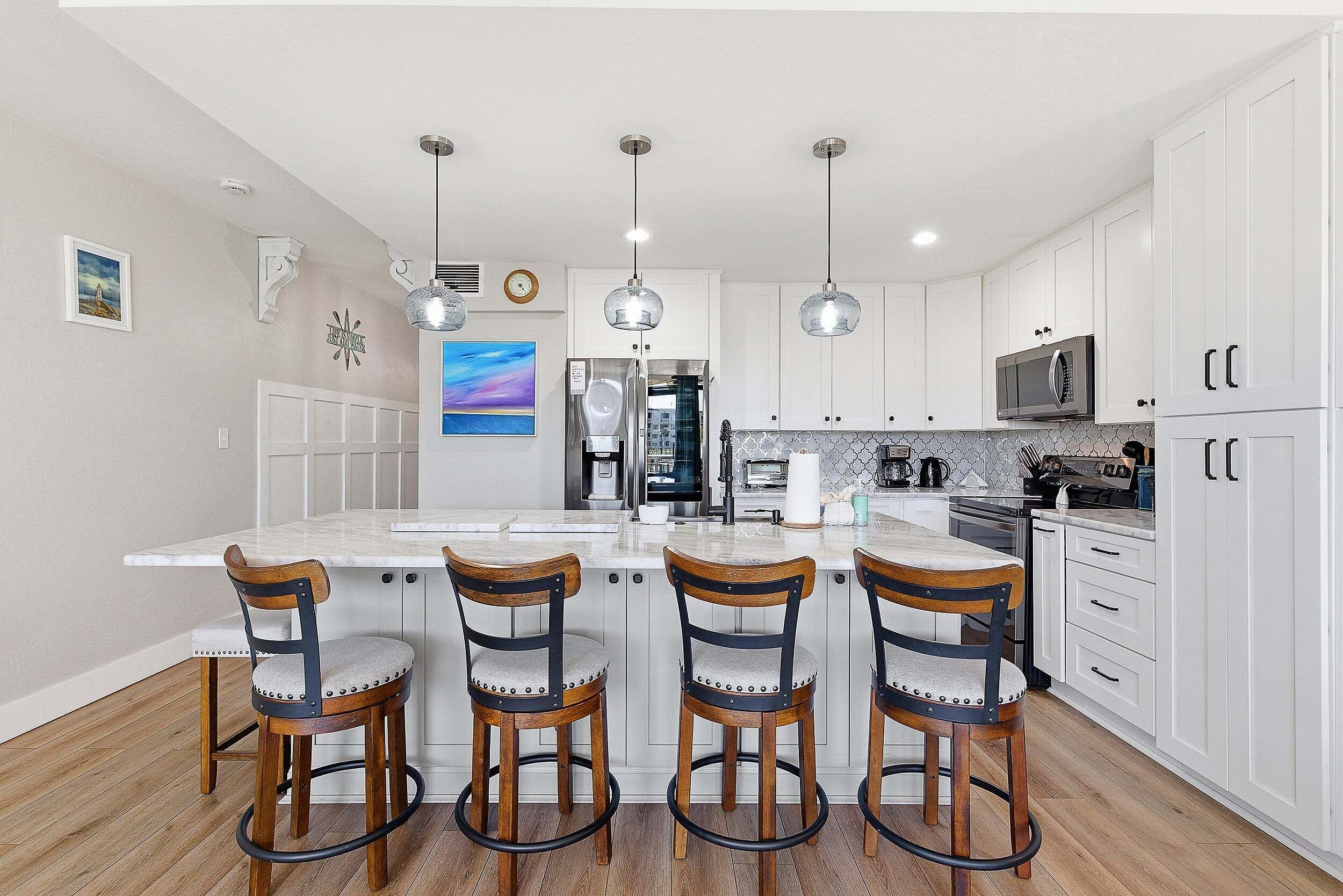 A modern kitchen with white cabinets, a central island with bar stools, stainless steel appliances, pendant lighting, and a view of the living area.