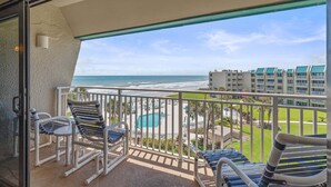 Un balcon avec vue sur la plage et la piscine, des chaises blanches et une porte vitrée.