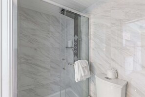 A glass-enclosed shower with a white towel hanging on a rack, a toilet with a tissue holder, and marble walls. - Hawaiian Inn 332 (Daytona Beach Shores)