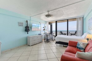 A room with a bed, a red couch, a guitar, a radiator, a table with chairs, and a view of the ocean. - Hawaiian Inn 332 (Daytona Beach Shores)