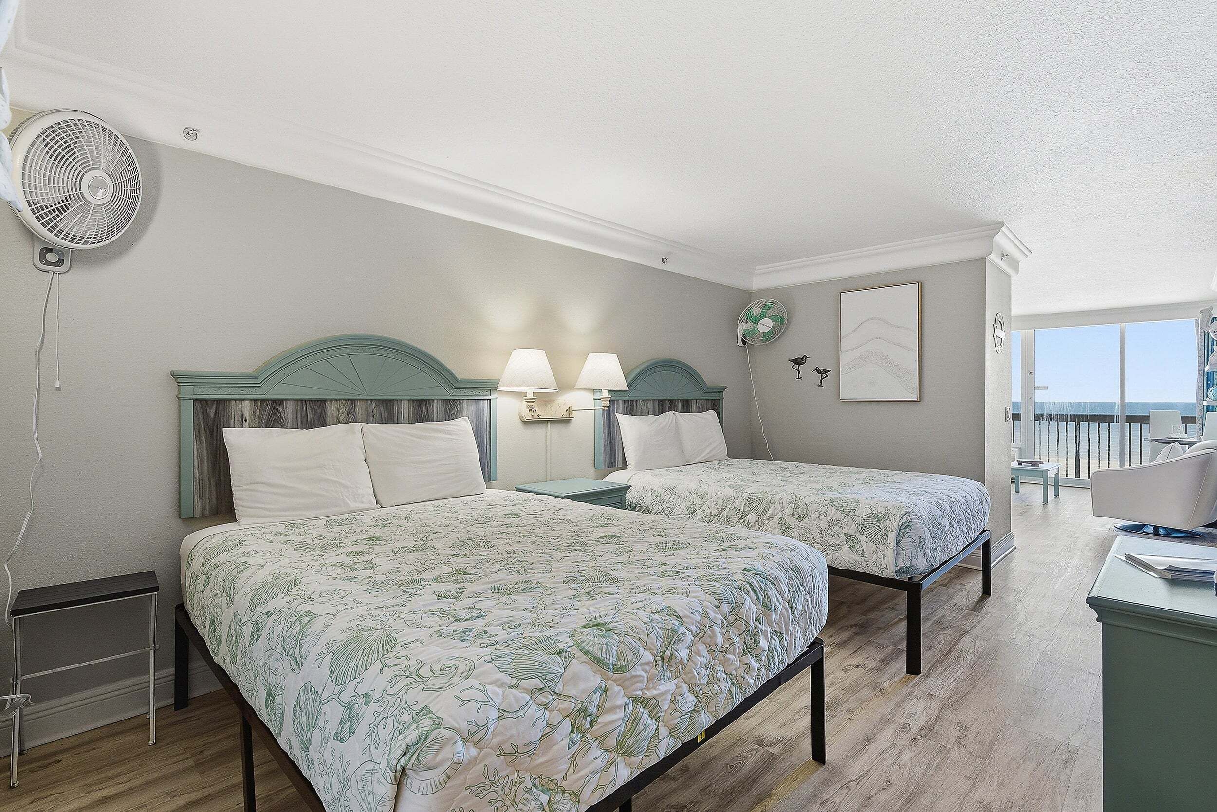 A bedroom with two beds, a standing fan, and a view of the balcony.