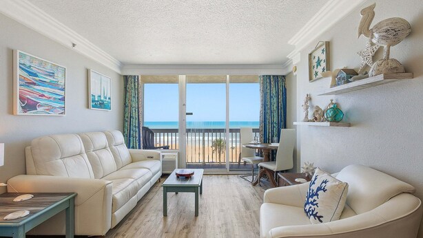 A living room with a sofa, armchair, coffee table, and a view of the beach through large windows. - Daytona Beach Resort 456 (Daytona Beach)