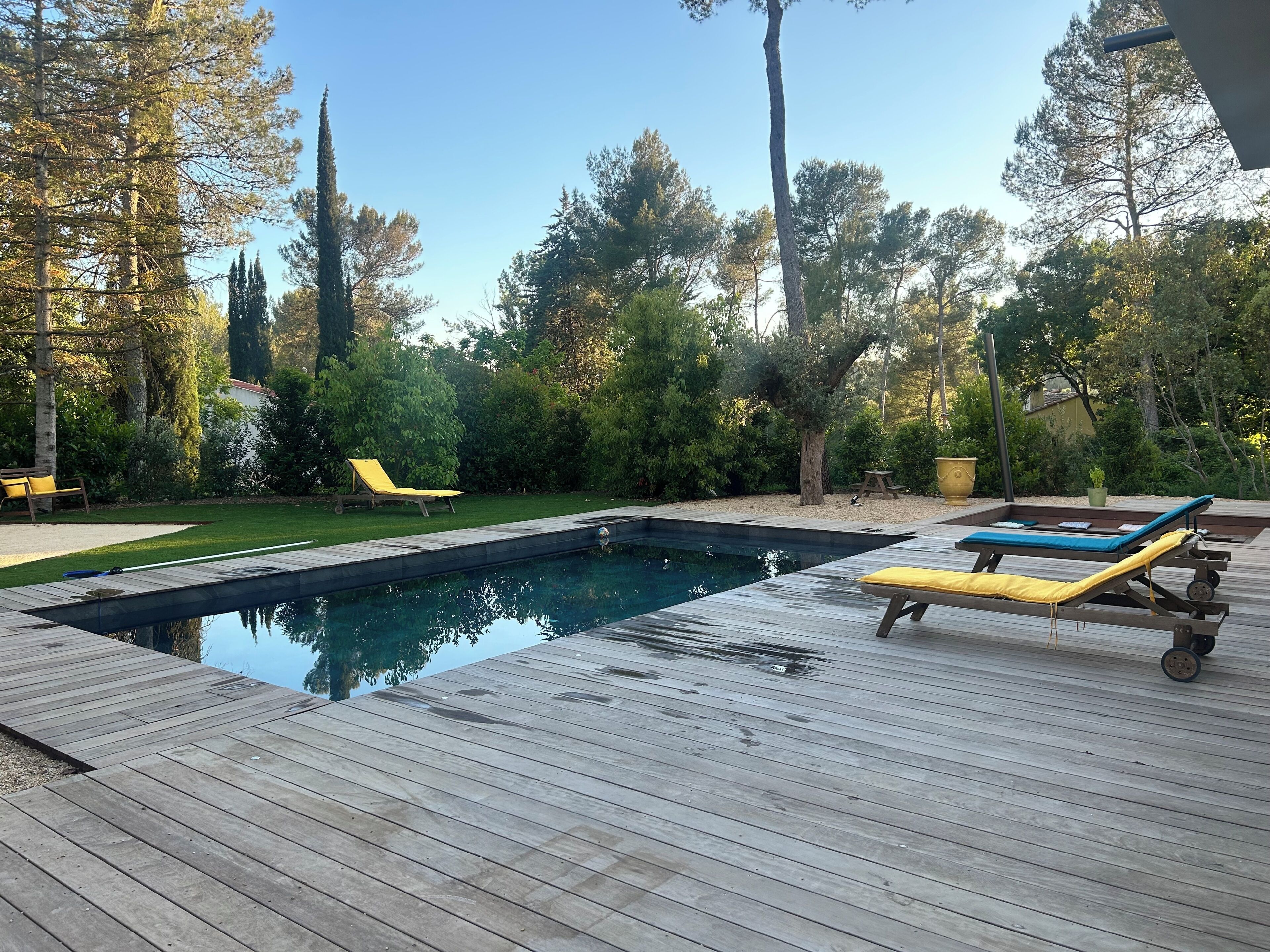 Une terrasse en bois avec une piscine, des chaises longues et un espace verdoyant agrémenté d’arbres.
