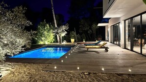 Une piscine avec des chaises longues, une terrasse en bois et une maison moderne dotée de grandes fenêtres.