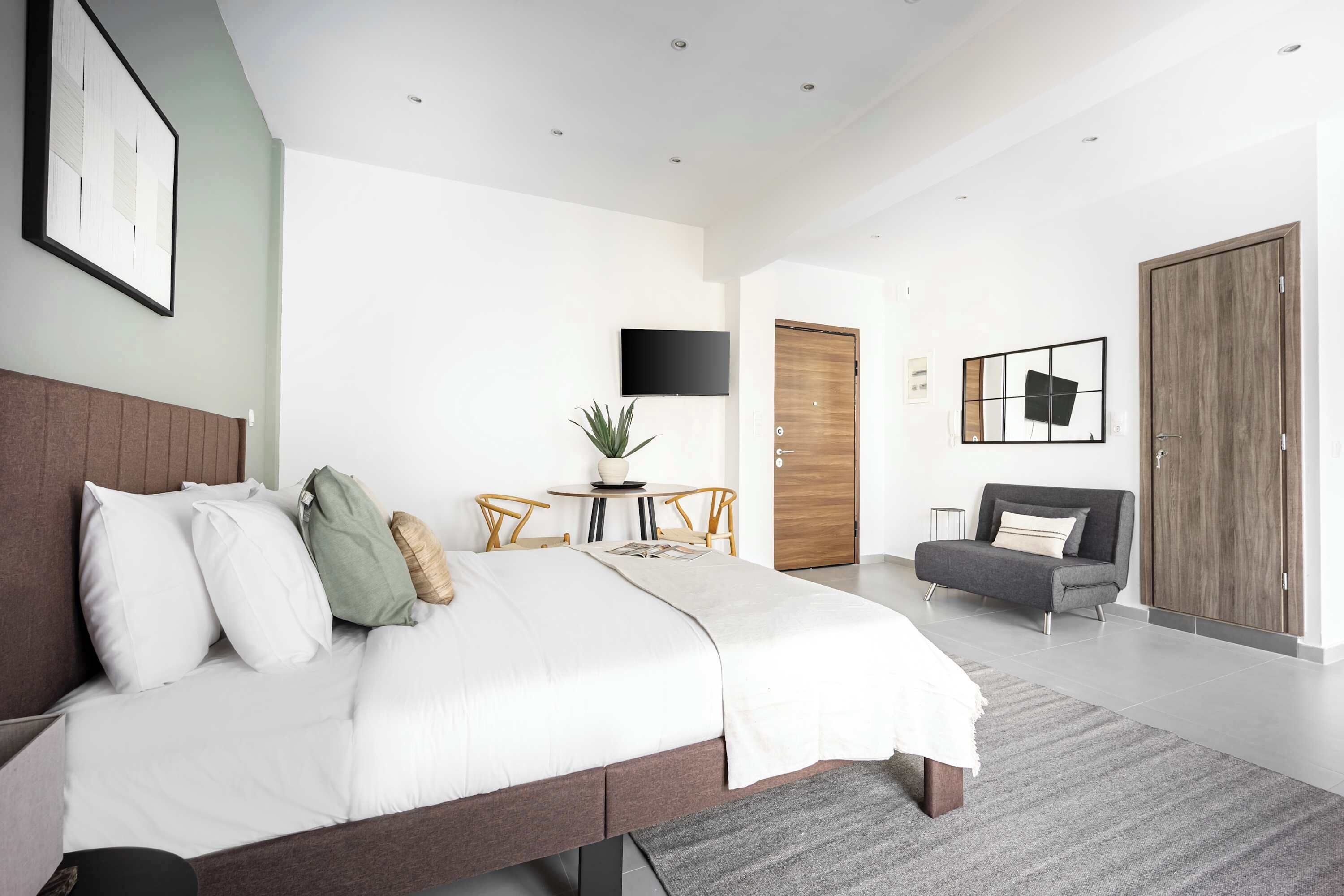 A modern bedroom with a large bed, a flat-screen TV, a small round table with chairs, a sofa, and a wardrobe.