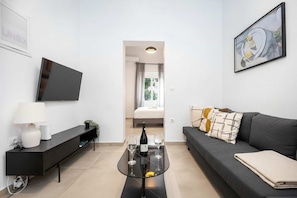 Un soggiorno moderno con una TV a schermo piatto, un tavolino in vetro con una bottiglia e bicchieri, un divano con cuscini decorati e un quadro appeso al muro.