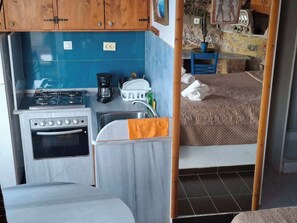 Eine kompakte Küchenzeile mit Kochfeld, Backofen und Spüle, ein Bett mit gesteppter Decke und ein kleiner Tisch mit einem Blumentopf.