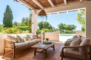 Ein überdachter Außenbereich mit einer Couch, Stühlen und einem Tisch, mit Blick auf einen Swimmingpool und üppiges Grün.