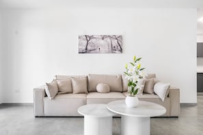 Ein modernes Wohnzimmer mit einem beigen Sofa, zwei runden Tischen, einer Zimmerpflanze und einem an der Wand hängenden Bild.