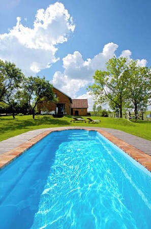 A swimming pool with a wooden deck, a brick house in the background, and a clear blue sky.