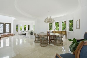 Ein geräumiges Wohnzimmer mit einem runden Holzesstisch, Korbstühlen, einer Pendelleuchte und großen Fenstern mit Blick ins Grüne.