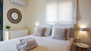Ein ordentlich bezogenes Bett mit Kissen, ein Nachttisch mit Lampe, eine Wand-Klimaanlage und ein dekorativer Spiegel.
