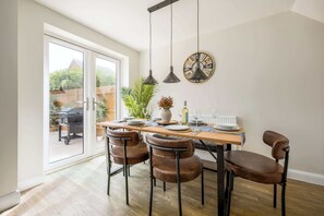 Ein Essbereich mit Holztisch, vier braunen Stühlen, hängenden Pendelleuchten, einer Wanduhr und Blick durch eine Glastür.