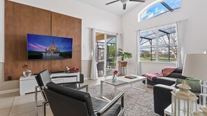 A spacious living room with a flat-screen TV, a ceiling fan, and a large window offering a view of the outdoors. - Lakefront 4BR Disney Home with Pool & Game Room (Kissimmee)