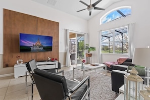 Ein geräumiges Wohnzimmer mit Flachbildfernseher, Deckenventilator und großem Fenster mit Blick ins Freie.