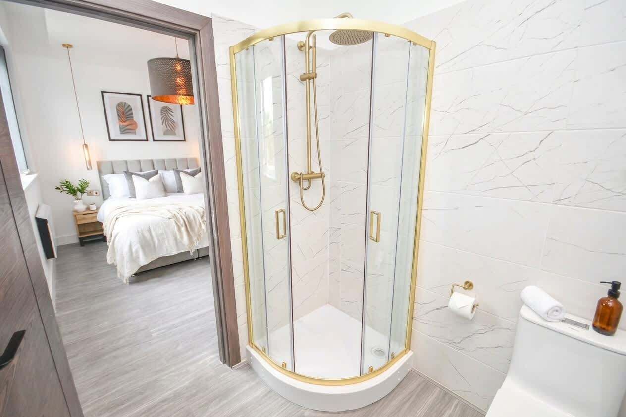 A modern bathroom with a glass-enclosed shower, a toilet, and a bed visible in the background.