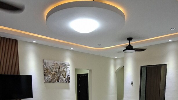 A spacious living area with a marble floor, a flat-screen TV mounted on a wooden panel, and a ceiling fan.